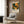 May include: Colorful art poster featuring abstract botanical design in orange, green, and cream tones with polka dots and organic shapes, displayed in light wood frame on wood-paneled wall above cream upholstered armchair with geometric patterned pillow, brass floor lamp and dark round side table with open book nearby