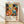 May include: Framed art print leaning against white wall on wooden floor with natural light shadows. Colorful abstract floral design with polka dots in vibrant oranges, teals, pinks, and navy blues. Text below reads "Yayoi Kusama" with Japanese characters and "Japan 1987". Light wood frame surrounds cream-colored poster with playful botanical patterns and circular motifs.