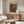 May include: Yayoi Kusama exhibition poster from 1992 displayed above a wooden sideboard in a minimalist living room. The poster features vibrant polka dot patterns in orange, purple, and yellow against a dark background with Japanese text. A white curved chair sits beside a glass-top coffee table, with modern lighting fixtures completing the mid-century modern aesthetic.