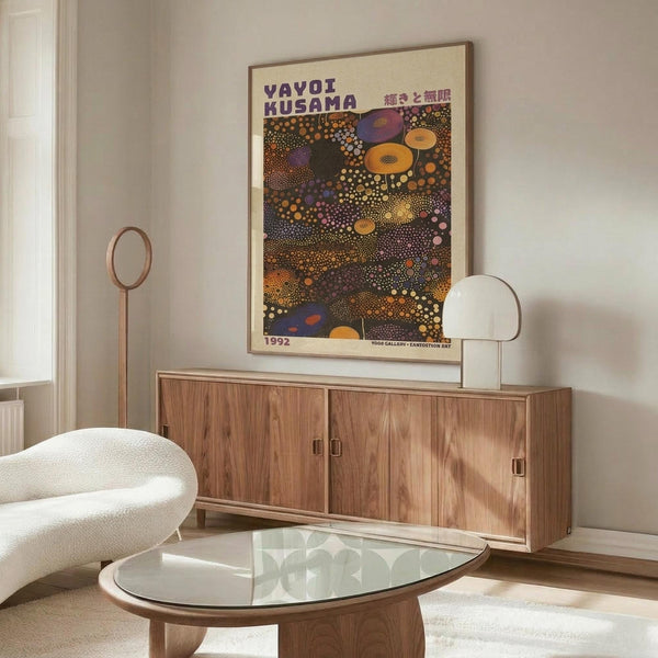 May include: Yayoi Kusama exhibition poster from 1992 displayed above a wooden sideboard in a minimalist living room. The poster features vibrant polka dot patterns in orange, purple, and yellow against a dark background with Japanese text. A white curved chair sits beside a glass-top coffee table, with modern lighting fixtures completing the mid-century modern aesthetic.