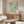 May include: Yayoi Kusama 1991 exhibition poster displayed above a wooden credenza in a modern living room. The poster features abstract teal and cream polka dot patterns in a cream frame. Room includes white sculptural chair, glass-top coffee table with wooden base, and minimalist table lamp. Natural lighting creates soft shadows on white walls.