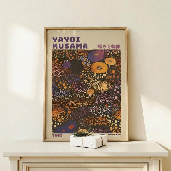 May include: Framed poster featuring colorful polka dot artwork in purple, orange, yellow and pink circles on dark background, displayed on light wood surface next to small wrapped gift box. The abstract design shows Kusama's signature infinity net and dot patterns in vibrant, overlapping circular forms creating a dreamlike composition.