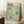 May include: Framed exhibition poster with cream background featuring abstract teal and turquoise dots, circles, and radiating patterns. Black text at top reads "YAYOI KUSAMA" with Japanese characters. Poster leans against white wall corner next to wooden side table with white coffee cup and saucer. Beige textured rug and neutral fabric visible in cozy interior setting with natural window lighting.
