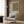 May include: Beige upholstered armchair with geometric patterned pillow positioned against wood-paneled wall. Framed poster shows olive green background with scattered white dots in polka dot pattern, Japanese text beneath. Gold floor lamp with white circular base stands nearby, open book rests on dark round side table. Neutral curtains and light wood flooring complete the modern minimalist room setting.