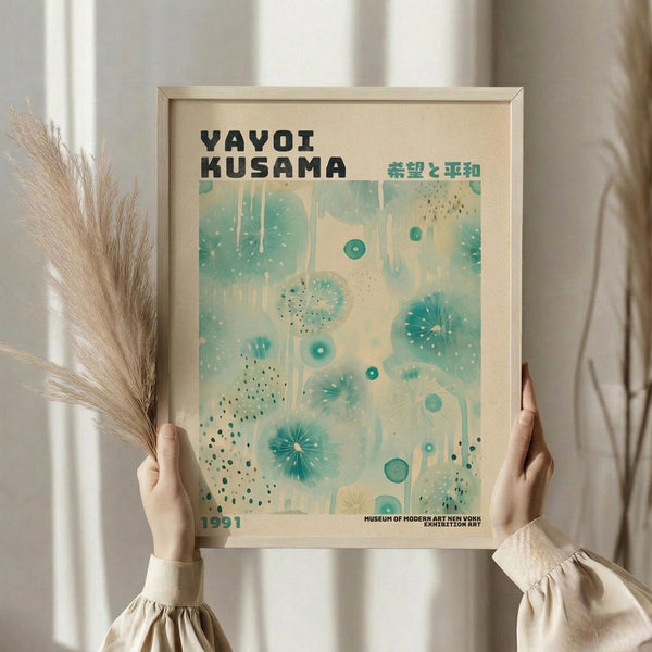 May include: Person in cream sweater holding framed vintage exhibition poster with teal abstract polka dot pattern against cream background, displayed alongside dried wheat stems in soft natural lighting
