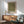 May include: Framed poster with olive green background and scattered white dots in various sizes, displayed above a wooden sideboard with white ceramic lamp. Mid-century modern living room setting features sage green upholstered chair with wooden arms, round coffee table, and white walls with natural sunlight creating shadows. The artwork shows Japanese text at bottom and has a light wooden frame.