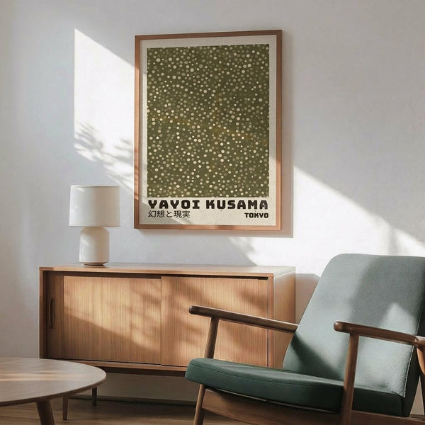 May include: Framed poster with olive green background and scattered white dots in various sizes, displayed above a wooden sideboard with white ceramic lamp. Mid-century modern living room setting features sage green upholstered chair with wooden arms, round coffee table, and white walls with natural sunlight creating shadows. The artwork shows Japanese text at bottom and has a light wooden frame.