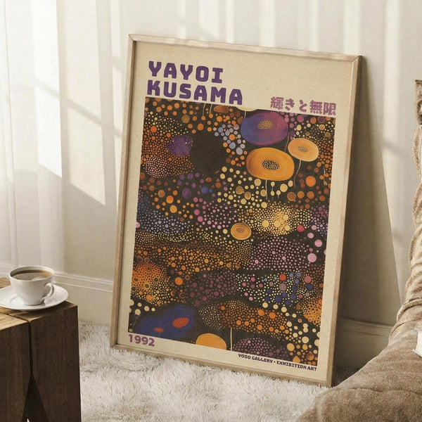 May include: Framed art poster leaning against white wall corner on fluffy white rug. Poster features colorful polka dot pattern with purple, orange, yellow and blue circles on dark background, resembling cosmic or cellular forms. Purple text at top with Japanese characters. Wooden frame, dated 1992. Coffee cup on wooden block nearby, beige blanket visible at right edge.