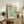May include: Framed vintage-style exhibition poster with turquoise and cream abstract dotted pattern, displayed on rustic wood sideboard alongside fern plant, stacked books, ceramic vase and table lamp in neutral-toned living room setting.