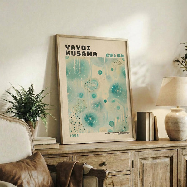 May include: Framed vintage-style exhibition poster with turquoise and cream abstract dotted pattern, displayed on rustic wood sideboard alongside fern plant, stacked books, ceramic vase and table lamp in neutral-toned living room setting.