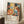 May include: Colorful abstract art poster in light wood frame leaning against white wall corner, featuring vibrant floral and polka dot patterns in yellow, orange, blue, and red with Japanese text at bottom. Small coffee cup on wooden block beside poster, cream knit blanket partially visible, white fluffy rug underneath.