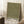 May include: Framed art print with olive green background covered in scattered cream and white polka dots of varying sizes, displayed in light wood frame leaning against white wall corner. Black text at bottom reads "YAYOI KUSAMA" with Japanese characters above and "TOKYO" below. Scene includes coffee cup on wooden block, textured white rug, and beige blanket, with natural window light creating geometric shadows on wall.