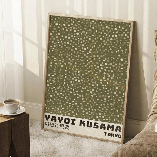 May include: Framed art print with olive green background covered in scattered cream and white polka dots of varying sizes, displayed in light wood frame leaning against white wall corner. Black text at bottom reads "YAYOI KUSAMA" with Japanese characters above and "TOKYO" below. Scene includes coffee cup on wooden block, textured white rug, and beige blanket, with natural window light creating geometric shadows on wall.