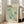 May include: Framed vintage-style exhibition poster with cream background and turquoise polka dot pattern, featuring artist name "YAYOI KUSAMA" in black text, Japanese characters, and "1991" date. The abstract design shows scattered circular dots and radiating burst patterns. White wooden frame leans against white wall next to beige upholstered sofa in bright, sunlit living room with tall windows.