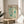 May include: Framed Yayoi Kusama exhibition poster featuring abstract turquoise and teal organic shapes, dots, and starburst patterns on cream background, displayed on light wood round side table next to beige upholstered chair with plant leaves casting shadows on white wall in bright natural lighting.