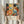 May include: Hands holding a framed colorful abstract floral art print featuring vibrant orange, yellow, and blue polka-dotted flowers and organic shapes. The poster displays "YAYOI KUSAMA" and "JAPAN 1987" text at bottom with Japanese characters. Dried pampas grass sits nearby against white curtains in soft natural lighting. The artwork shows Kusama's signature dotted style with bold botanical motifs in warm and cool tones within a light wood frame.