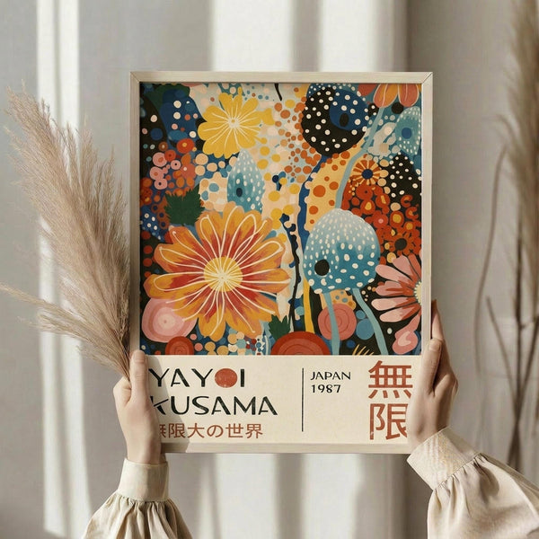 May include: Hands holding a framed colorful abstract floral art print featuring vibrant orange, yellow, and blue polka-dotted flowers and organic shapes. The poster displays "YAYOI KUSAMA" and "JAPAN 1987" text at bottom with Japanese characters. Dried pampas grass sits nearby against white curtains in soft natural lighting. The artwork shows Kusama's signature dotted style with bold botanical motifs in warm and cool tones within a light wood frame.