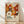 May include: Framed poster print leaning against white wall on light wood floor. Artwork features abstract floral pattern in warm oranges, browns, and cream tones with polka dots and radiating flower shapes. Text shows "YAYOI KUSAMA" with Japanese characters and "Musée d'Art Moderne de la Ville de Paris" in thin wooden frame. Soft natural lighting creates diagonal shadows across white fabric backdrop.