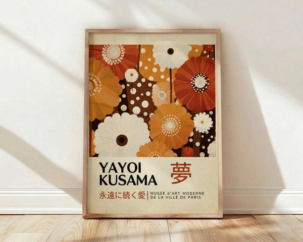 May include: Framed poster print leaning against white wall on light wood floor. Artwork features abstract floral pattern in warm oranges, browns, and cream tones with polka dots and radiating flower shapes. Text shows "YAYOI KUSAMA" with Japanese characters and "Musée d'Art Moderne de la Ville de Paris" in thin wooden frame. Soft natural lighting creates diagonal shadows across white fabric backdrop.