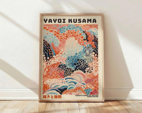 May include: Framed poster with abstract dotted pattern in coral, blue, and black colors featuring flowing organic shapes and wave-like elements at bottom. Text at top reads "YAYOI KUSAMA" with Japanese characters below. Wood frame sits on light wooden floor against white wall with natural lighting casting soft shadows.