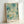 May include: Framed art print with abstract aqua and teal watercolor background featuring scattered circles and dots in various sizes, vertical dripping lines, and Japanese text at bottom. Light wooden frame leaning against white wall on light surface.