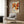 May include: A framed poster featuring abstract floral patterns in warm orange, brown, and cream tones with polka dot elements and Japanese text reading "Yayoi Kusama." The artwork is displayed on a wood-paneled wall above a beige upholstered chair with geometric patterned pillow. A brass floor lamp and dark round side table with open book complete the modern living room setting with natural lighting from nearby curtained window.