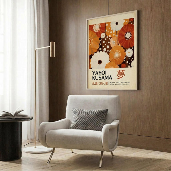 May include: A framed poster featuring abstract floral patterns in warm orange, brown, and cream tones with polka dot elements and Japanese text reading "Yayoi Kusama." The artwork is displayed on a wood-paneled wall above a beige upholstered chair with geometric patterned pillow. A brass floor lamp and dark round side table with open book complete the modern living room setting with natural lighting from nearby curtained window.
