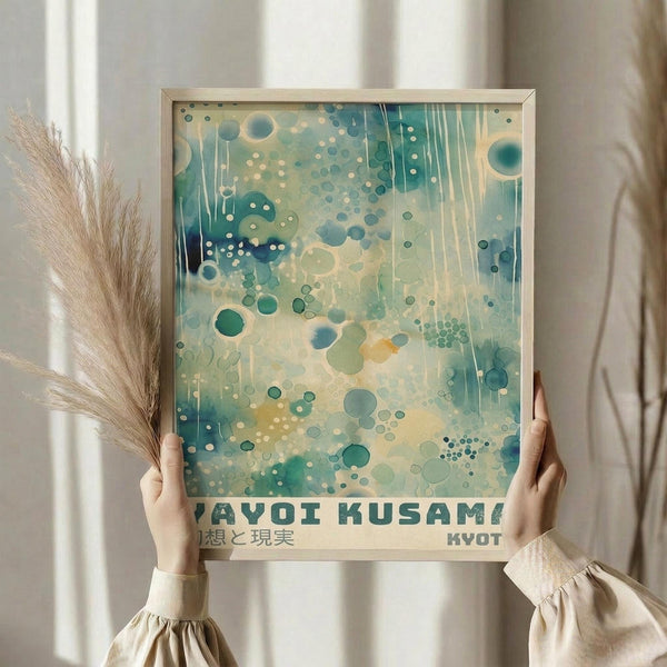 May include: Person holding framed abstract art print with teal and cream watercolor circles, dots, and vertical streaks. Text reads "YAYOI KUSAMA" and Japanese characters below, with "KYOTO" in corner. Natural wood frame, beige clothing, dried pampas grass, soft lighting near white curtained window.