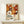 May include: Framed poster with abstract floral pattern in warm orange, brown, and cream colors featuring circular flower motifs and polka dots. Text reads "YAYOI KUSAMA" with Japanese characters and "MUSEE d'ART MODERNE DE LA VILLE DE PARIS" below. Light wood frame sits on white wooden furniture beside small wrapped gift with greenery. Neutral beige wall background creates minimalist display setting.
