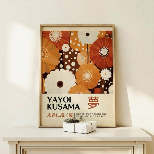 May include: Framed poster with abstract floral pattern in warm orange, brown, and cream colors featuring circular flower motifs and polka dots. Text reads "YAYOI KUSAMA" with Japanese characters and "MUSEE d'ART MODERNE DE LA VILLE DE PARIS" below. Light wood frame sits on white wooden furniture beside small wrapped gift with greenery. Neutral beige wall background creates minimalist display setting.
