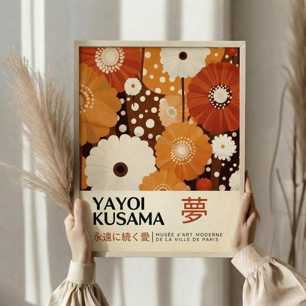 May include: Hands holding a framed art print featuring retro-style floral patterns in warm orange, cream, and brown tones with polka dot accents. Text reads "Yayoi Kusama" with Japanese characters and "Musée d'Art Moderne de la Ville de Paris." Light wood frame against neutral curtained background with dried pampas grass visible on left side.