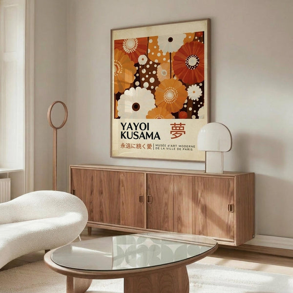 May include: Framed exhibition poster featuring colorful geometric flowers in warm orange, brown, and cream tones displayed above a mid-century modern walnut credenza. Modern living room setting with white curved chair, glass oval coffee table, and brass floor lamp on light wood floors against neutral walls.