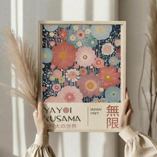 May include: Hands holding a framed art print featuring colorful abstract floral design with coral, pink, and blue blooms against a dark navy background. The poster has "YAYOI KUSAMA" and "JAPAN 1987" text at bottom with Japanese characters. Light wood frame, beige curtains and dried pampas grass visible in soft, natural lighting.