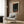 May include: Colorful poster with cosmic, swirling design featuring orange and red dots against dark background, displayed in light wood frame on wood-paneled wall. Modern reading corner with cream upholstered armchair, geometric patterned pillow, brass floor lamp, and dark round side table with open book. Warm neutral tones throughout contemporary interior space.