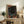 May include: Framed poster with abstract cosmic design featuring swirling patterns in dark background with orange and red circular dots, displayed on rustic wooden console table alongside potted fern, books, and ceramic vase in neutral-toned living room setting with beige armchair visible.