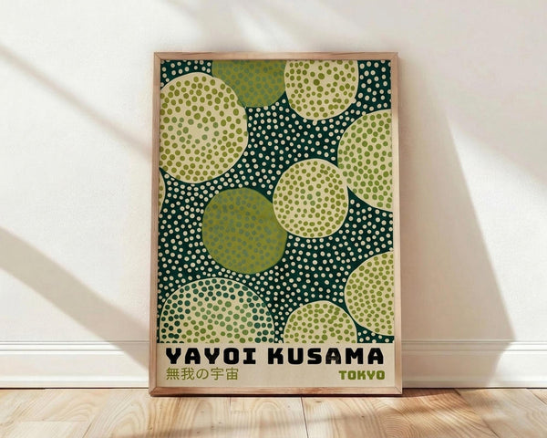 May include: Framed poster print leaning against white wall on light wood floor. Artwork features abstract pattern of overlapping circles in various shades of green - from dark forest to lime green - filled with polka dots in white and darker green. Text reads "YAYOI KUSAMA" with Japanese characters and "TOKYO" below. Natural wood frame with visible grain. Soft natural lighting creates gentle shadows on white fabric backdrop.