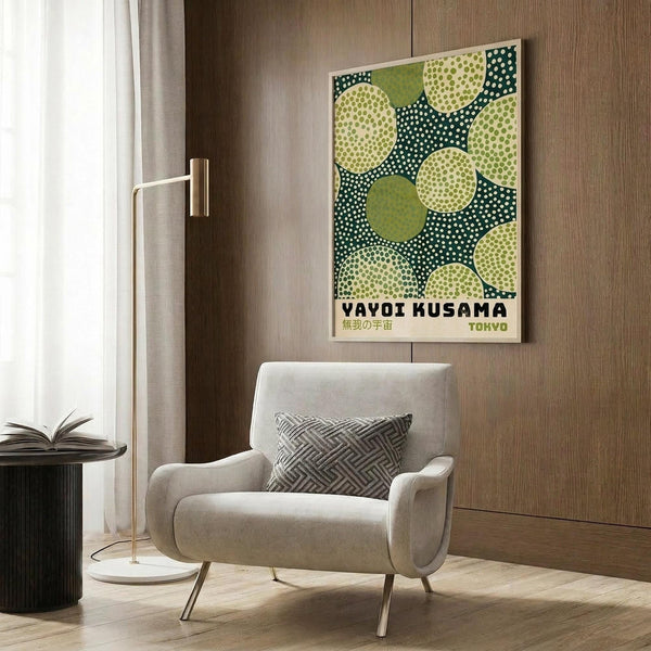 May include: Modern beige armchair with geometric pillow in contemporary living room setting. Framed poster on wood paneled wall features green polka dot pattern with circular motifs and Japanese text. Gold floor lamp and dark round side table with open book visible. Warm wood tones and neutral textiles create minimalist interior design aesthetic.