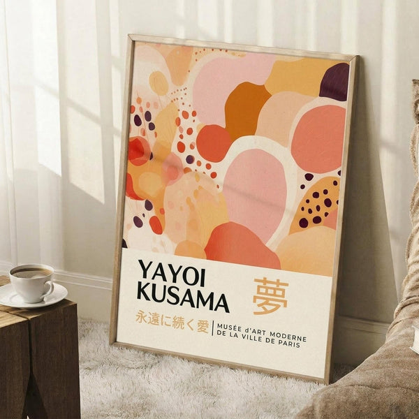May include: Framed abstract art poster with flowing organic shapes in warm peach, coral, orange, and burgundy tones with scattered dots, displayed in light wood frame leaning against white wall. Scene shows white coffee cup on wooden block, cream textured rug, and beige blanket. Text reads "Yayoi Kusama" with Japanese characters and "Musee d'Art Moderne de la Ville de Paris" at bottom.