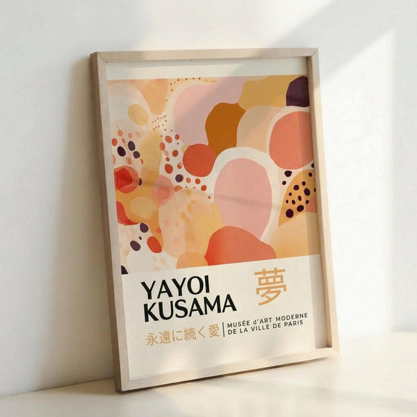 May include: Abstract art poster in light wood frame leaning against white wall. Artwork features flowing organic shapes in warm coral, orange, pink, yellow and burgundy tones with scattered dots throughout. Text reads "YAYOI KUSAMA" in black letters with Japanese characters and "MUSÉE D'ART MODERNE DE LA VILLE DE PARIS" below on cream background.