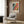 May include: Framed Yayoi Kusama exhibition poster displayed on wood-paneled wall in modern living room with beige armchair, floor lamp, and side table. Colorful abstract artwork features flowing bands of pink, orange, blue, and black with polka dots. White frame complements neutral decor with geometric throw pillow and hardwood flooring.