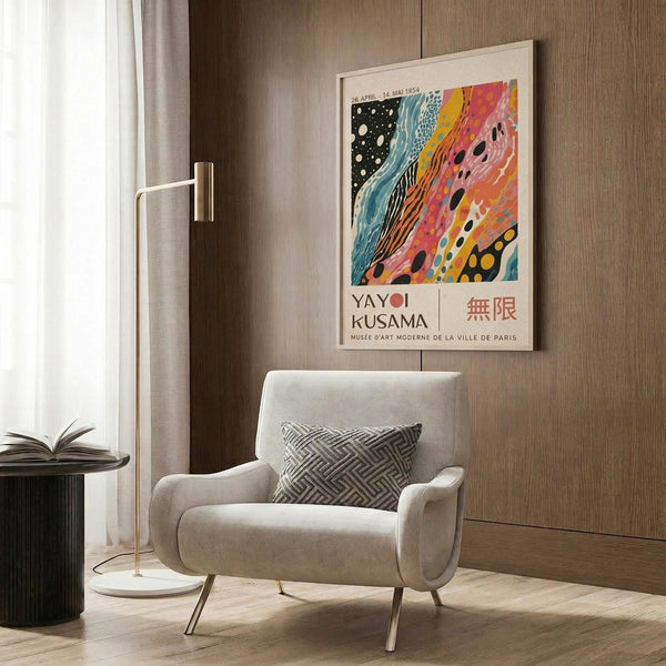 May include: Framed Yayoi Kusama exhibition poster displayed on wood-paneled wall in modern living room with beige armchair, floor lamp, and side table. Colorful abstract artwork features flowing bands of pink, orange, blue, and black with polka dots. White frame complements neutral decor with geometric throw pillow and hardwood flooring.