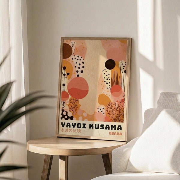 May include: Modern art poster in wooden frame displayed on round wooden side table next to beige armchair and plant. Abstract artwork features warm coral, orange, brown and cream organic shapes, dots, and botanical elements. Japanese text at bottom. Soft natural lighting creates shadows on white wall.