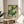 May include: Framed poster with green and cream polka dot pattern featuring overlapping circles in various shades of green against a dark teal background with white dots. Text reads "Yayoi Kusama" and "Tokyo" in black lettering. Wooden frame sits on light wood round side table next to beige upholstered chair. Natural sunlight streams across white walls, with plant leaves visible in corner creating soft shadows.