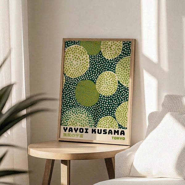 May include: Framed poster with green and cream polka dot pattern featuring overlapping circles in various shades of green against a dark teal background with white dots. Text reads "Yayoi Kusama" and "Tokyo" in black lettering. Wooden frame sits on light wood round side table next to beige upholstered chair. Natural sunlight streams across white walls, with plant leaves visible in corner creating soft shadows.