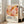 May include: A framed poster featuring abstract art with overlapping organic shapes in warm tones of coral, peach, orange, and burgundy, decorated with scattered dots. The poster displays "YAYOI KUSAMA" in black text with Japanese characters and museum attribution below. The light wooden frame leans against a white wall next to a beige upholstered sofa in a bright, minimalist living room with large windows.