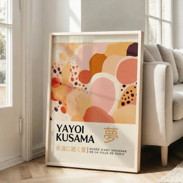 May include: A framed poster featuring abstract art with overlapping organic shapes in warm tones of coral, peach, orange, and burgundy, decorated with scattered dots. The poster displays "YAYOI KUSAMA" in black text with Japanese characters and museum attribution below. The light wooden frame leans against a white wall next to a beige upholstered sofa in a bright, minimalist living room with large windows.