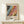 May include: Framed exhibition poster with abstract colorful artwork featuring flowing patterns in black, blue, pink, yellow and orange tones, displayed on light wood surface with small white gift box beside it. Natural lighting creates soft shadows on cream wall background.