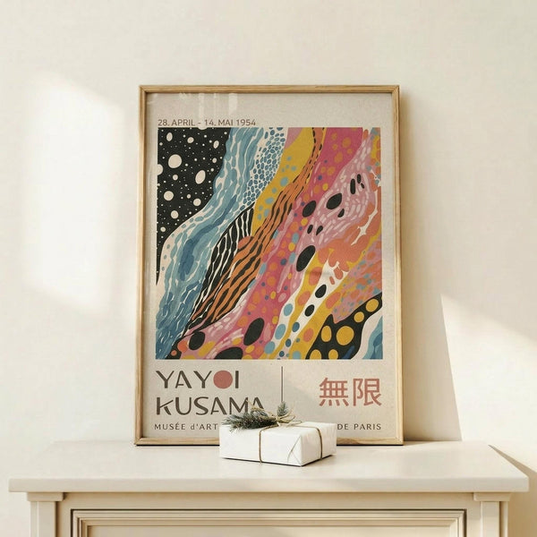 May include: Framed exhibition poster with abstract colorful artwork featuring flowing patterns in black, blue, pink, yellow and orange tones, displayed on light wood surface with small white gift box beside it. Natural lighting creates soft shadows on cream wall background.