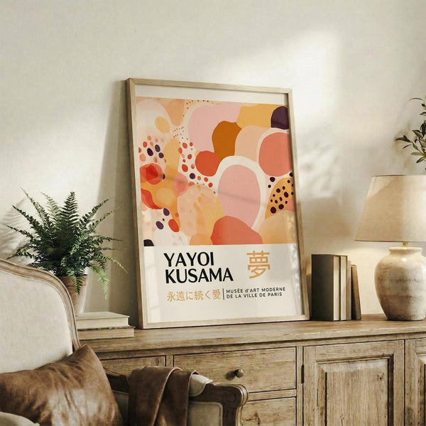 May include: Framed abstract art print with warm orange, pink, and coral organic shapes and dotted patterns, displayed on rustic wooden sideboard alongside fern plant, ceramic vase, and leather chair in neutral-toned living room setting
