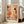 May include: Framed abstract art print leaning against white wall beside beige sofa in bright living room. Artwork features warm coral, orange, and brown organic shapes, dots, and botanical elements on cream background. Text reads "YAYOI KUSAMA" with Japanese characters and "OSAKA" below. Light wood frame, approximately 2-3 feet tall, positioned on light wooden floor near large windows.